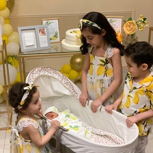 Op de afbeelding: Een pasgeboren baby in een wieg, omringd door drie kinderen. De baby draagt een outfit met citroenthema. De kinderen dragen bijpassende jurken en shirts met citroenprint. De sc&egrave;ne is versierd met gele ballonnen en een taart.
