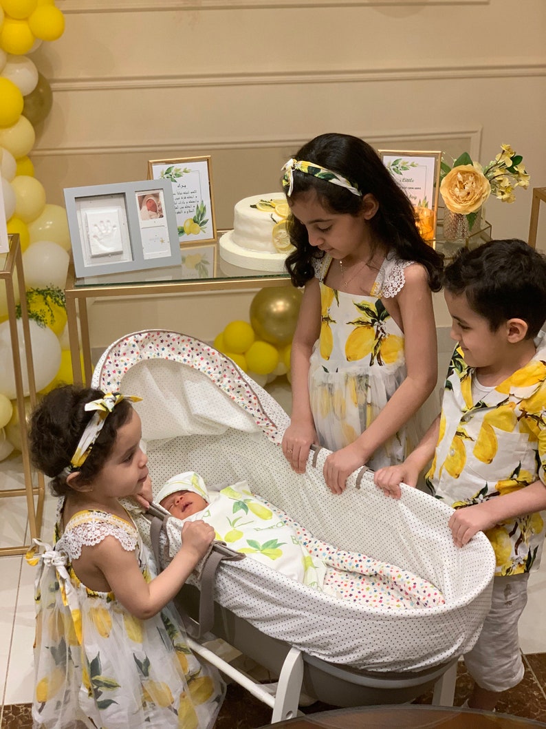Op de afbeelding: Een pasgeboren baby in een wieg, omringd door drie kinderen. De baby draagt een outfit met citroenthema. De kinderen dragen bijpassende jurken en shirts met citroenprint. De sc&egrave;ne is versierd met gele ballonnen en een taart.