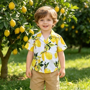 Op de afbeelding: Een kind draagt een shirt met citroenprint en een kaki korte broek. Het witte shirt is bedrukt met gele citroenen en groene bladeren. De achtergrond toont een citroenboom, wat een zomers tafereel cre&euml;ert.