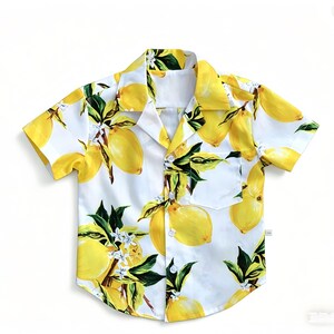 Op de afbeelding: Een shirt met korte mouwen met een citroenprint. Het shirt heeft een witte achtergrond versierd met gele citroenen, groene bladeren en kleine witte bloemen. Het heeft een borstzak en een klassieke kraag.