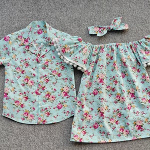 Puede incluir: Conjunto a juego de ropa infantil con estampado floral. El conjunto incluye una camisa de manga corta, un vestido con hombros descubiertos y una diadema. La tela es azul claro con flores rosas y amarillas.