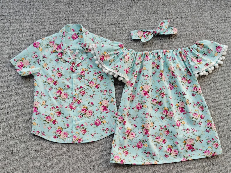 Puede incluir: Conjunto a juego de ropa infantil con estampado floral. El conjunto incluye una camisa de manga corta, un vestido con hombros descubiertos y una diadema. La tela es azul claro con flores rosas y amarillas.