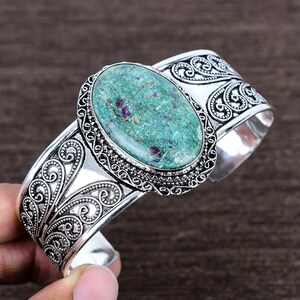 May include: Silver cuff bracelet featuring an oval, green gemstone with red inclusions. The gemstone is set in a detailed, ornate silver frame. The bracelet has an open design with intricate scrollwork patterns.