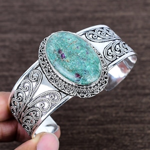May include: A silver cuff bracelet with an oval, green stone centre. The stone has red flecks and is set in a detailed, ornate silver frame. The bracelet features intricate scrollwork and a polished finish.