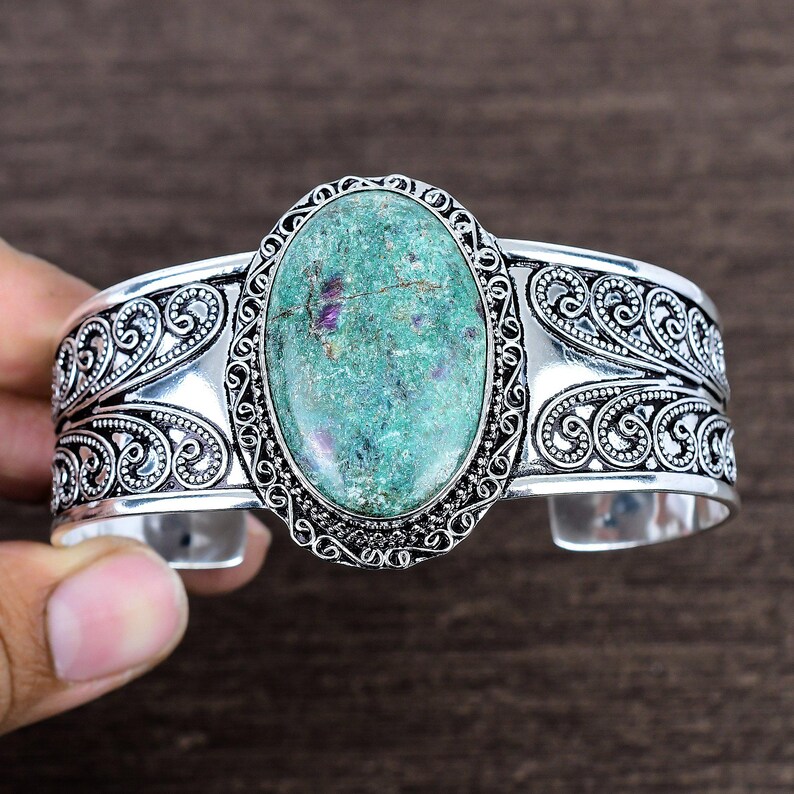 May include: Silver cuff bracelet featuring an oval, turquoise-colored stone with purple inclusions. The bracelet has an ornate, filigree design around the stone and along the band. The cuff is held by a hand.