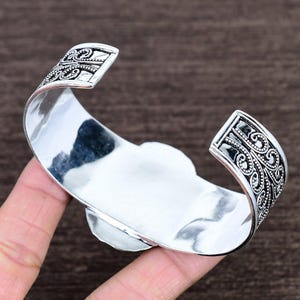 May include: A wide, open silver cuff bracelet with ornate scrollwork on the ends. The bracelet has a polished, reflective surface and a central, light-colored stone. The design is bold and eye-catching, suitable for everyday wear or special occasions.