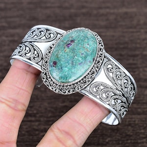 May include: Silver cuff bracelet featuring an oval, turquoise-colored stone with red inclusions. The bracelet has an ornate, filigree design with scrollwork and a detailed border around the stone. A unique piece of jewelry.