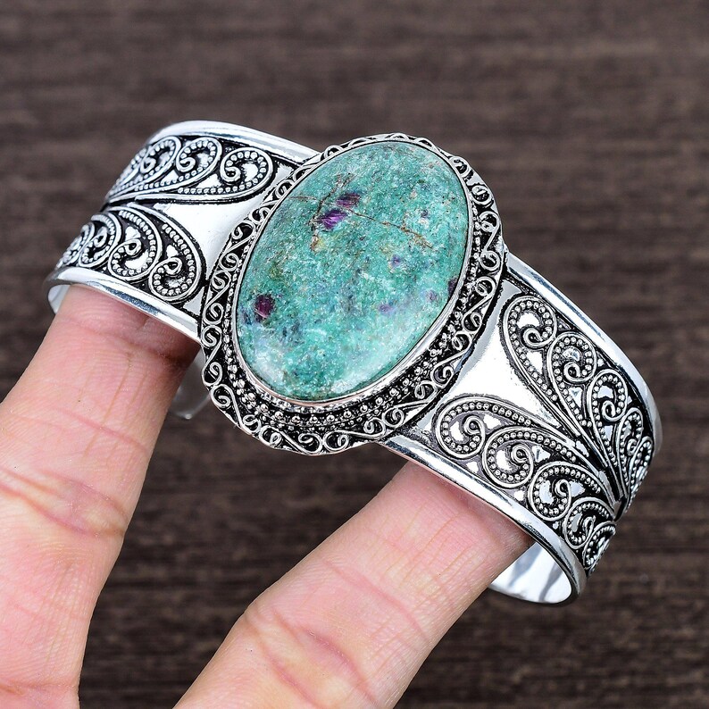 May include: Silver cuff bracelet featuring an oval, turquoise-colored stone with red inclusions. The bracelet has an ornate, filigree design with scrollwork and a detailed border around the stone. A unique piece of jewelry.