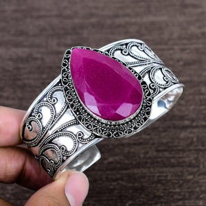 Op de afbeelding: Een zilveren manchetarmband met een sierlijk patroon, gezet met een grote, druppelvormige, magenta edelsteen. De armband heeft gedetailleerd krulwerk en een gepolijste afwerking, die vintage en modern combineert.