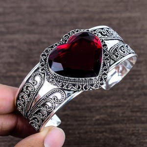 Puede incluir: Una pulsera de plata con un gran corazón de piedra preciosa roja. La pulsera tiene intrincados detalles de volutas ornamentales alrededor de la piedra preciosa y a lo largo de la banda. El diseño abierto permite un uso fácil.