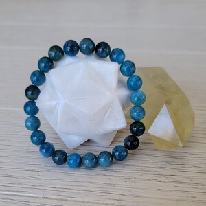 May include: A blue apatite gemstone bracelet with round beads. The bracelet is displayed on a white star-shaped object and a yellow crystal.