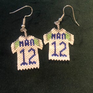 May include: A pair of white beaded earrings shaped like a soccer jersey with the number 12 and the word "SEA" on the front. The jersey is trimmed in green and blue.