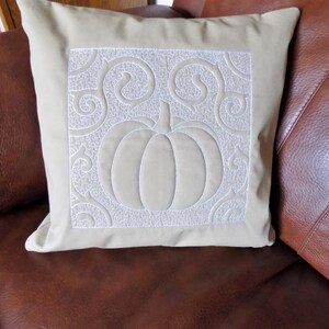 May include: A beige cushion with a white embroidered pumpkin and swirling vine design.