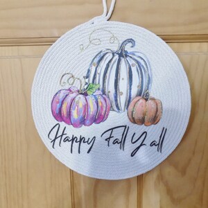 May include: A round, white, rope-wrapped door hanger with a painted design of three pumpkins. The pumpkins are painted in shades of pink, orange, and white with black stripes. The text "Happy Fall Y'all" is written in black below the pumpkins.