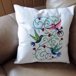 May include: White pillow with embroidered hummingbirds in shades of green, blue, pink, and purple, surrounded by green vines and red flowers.