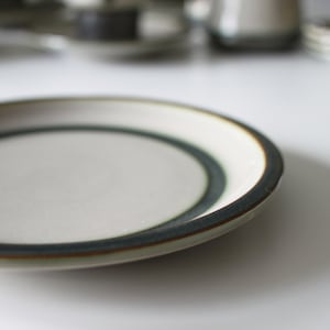 May include: A white ceramic saucer with a green and brown rim. The saucer is empty and is sitting on a white surface.