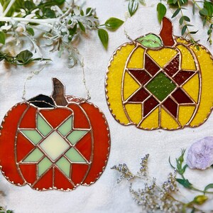 Peut inclure: Deux décorations de citrouille en vitrail avec un motif en étoile. La citrouille de gauche est rouge avec une étoile verte et blanche. La citrouille de droite est jaune avec une étoile rouge, verte et marron.