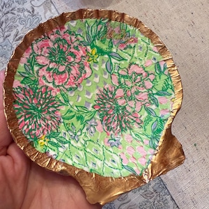 May include: A decorative seashell dish with a gold-painted rim. The interior features a floral pattern in pink, green, and white. The dish is designed to hold small items and adds a touch of coastal charm.