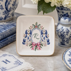 May include: A square white ceramic trinket dish with a gold-trimmed edge. The center features a decorative illustration of two blue and white spaniel dogs flanking a central crest containing the letter R, accented by holly leaves, red berries, and a pink bow.