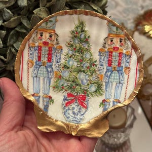 May include: A decorative seashell featuring a Christmas theme. The shell is adorned with a festive design of two nutcrackers, a Christmas tree, and a blue and white vase with a red bow. The shell's edge is trimmed with gold.