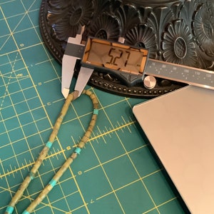 May include: A green and turquoise beaded necklace measured with a digital caliper reading 5.27 inches. The necklace is lying on a green cutting mat.