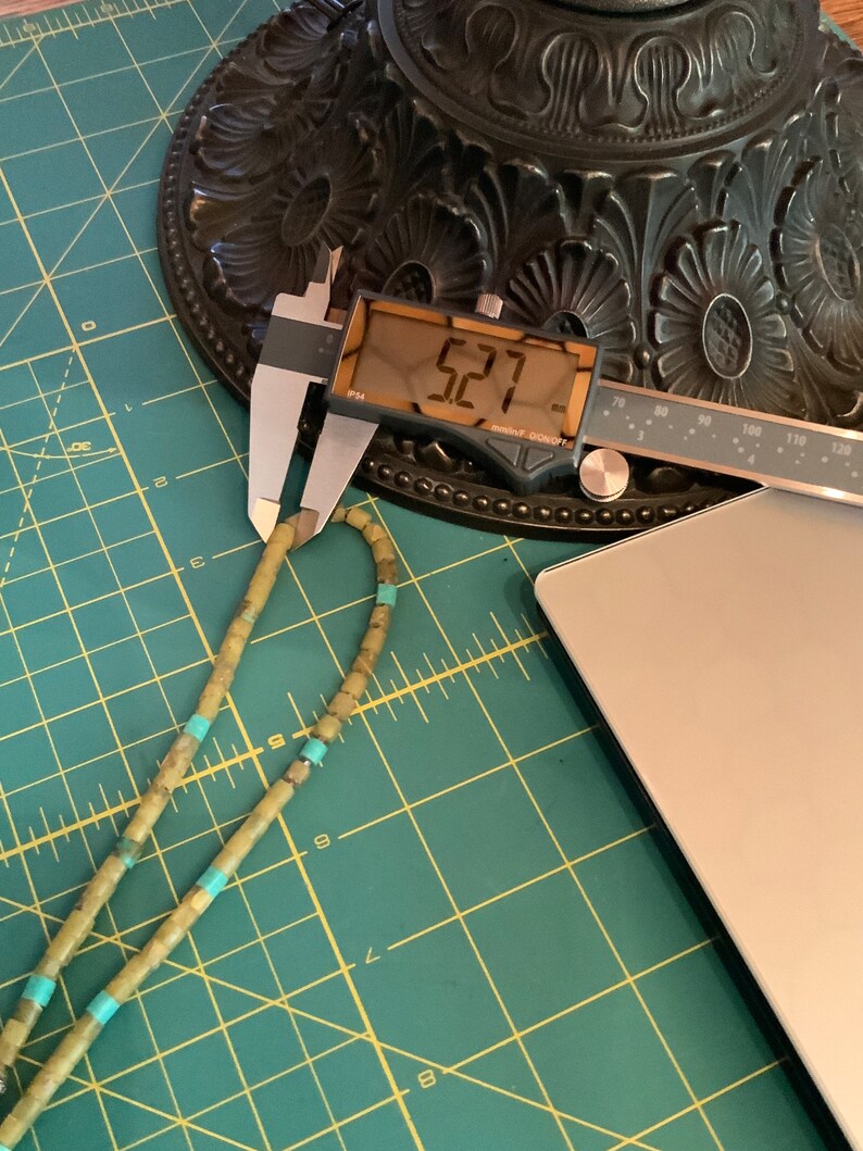May include: A green and turquoise beaded necklace measured with a digital caliper reading 5.27 inches. The necklace is lying on a green cutting mat.