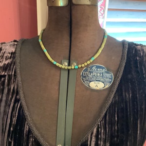 May include: A brown velvet dress with a green and blue beaded necklace. The dress has a V-neckline and a tie at the front. The dress is on a dress form. The dress form has a label that says "Acme Lullapsible Dress Form" and "Size A".
