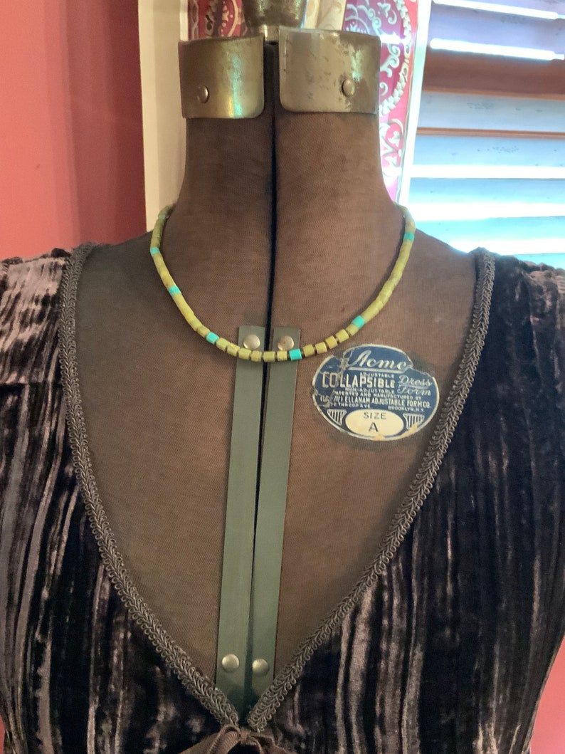 May include: A brown velvet dress with a green and blue beaded necklace. The dress has a V-neckline and a tie at the front. The dress is on a dress form. The dress form has a label that says "Acme Lullapsible Dress Form" and "Size A".