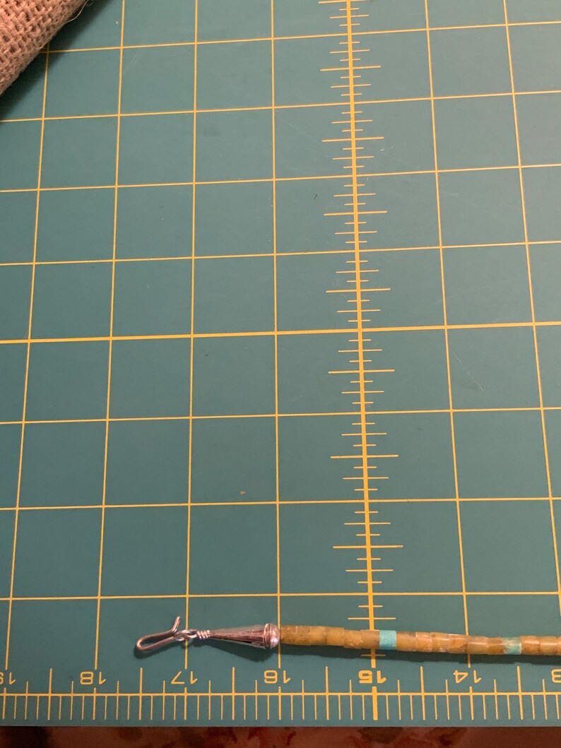 May include: A close-up of a turquoise and green beaded necklace with a silver clasp. The necklace is lying on a green cutting mat with white markings.