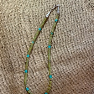 May include: A green and turquoise beaded necklace with silver clasps. The necklace is made of square beads and is strung on a cord.