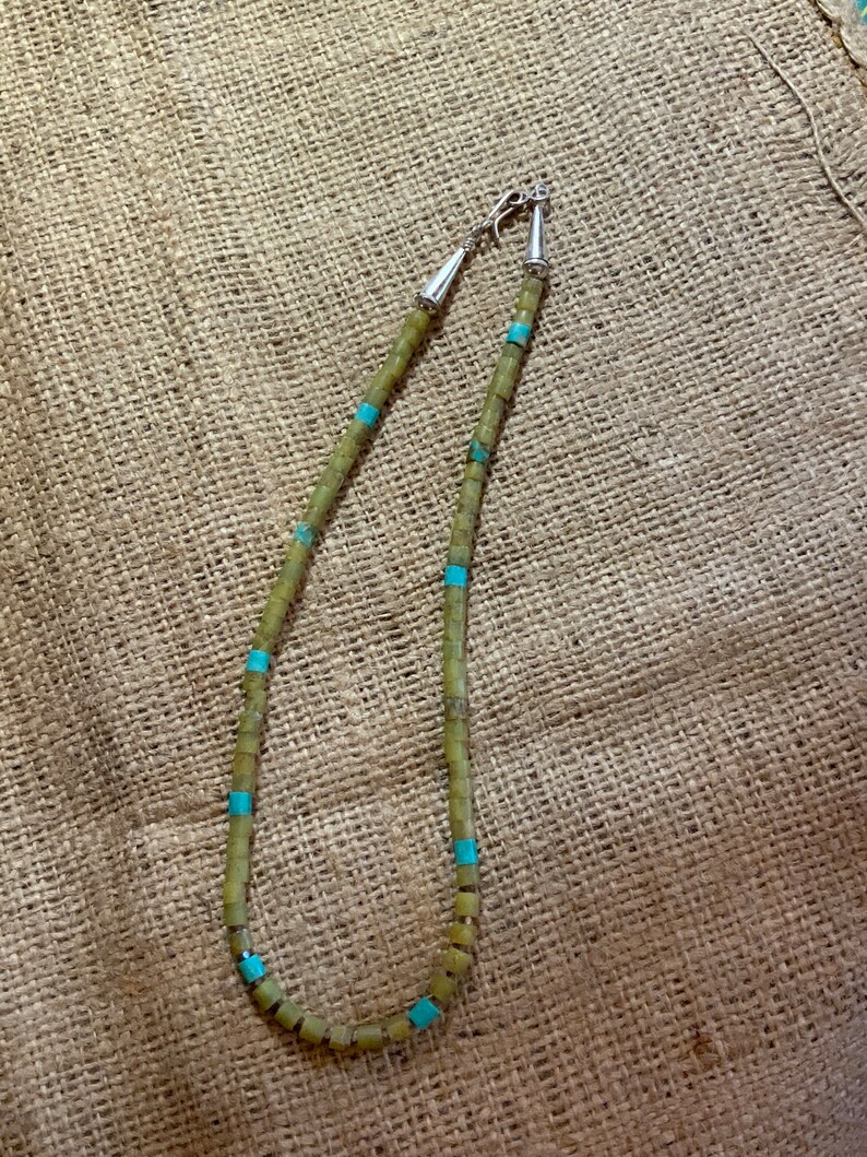 May include: A green and turquoise beaded necklace with silver clasps. The necklace is made of square beads and is strung on a cord.