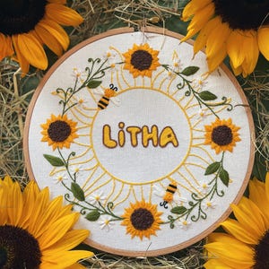 以下が含まれることがあります： 黄色で「Litha」という名前が刺繍されたフープアート。ひまわり、デイジー、ミツバチが周りに描かれています。デザインは白い布に木製のフープで施されています。明るい黄色のひまわりが作品を囲んでいます。