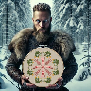 May include: A man with a beard holds a circular embroidery hoop with a white background. The embroidery features a red and yellow Vegvisir symbol surrounded by green and yellow leaves.