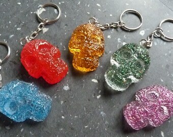 May include: Six colorful skull keychains with silver key rings. The skulls are made of clear plastic and have different colors and textures, including blue, red, green, orange, and pink.