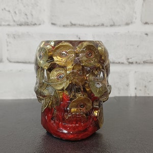 May include: A red skull-shaped shot glass with gold bullet casings embedded in the resin. The casings are visible through the clear resin.