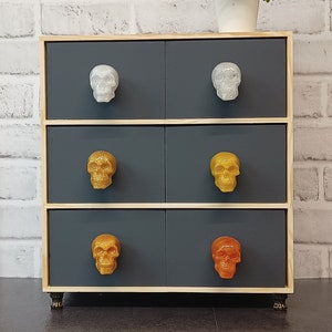 May include: A four-drawer chest of drawers with a light wood frame and dark grey drawers. Each drawer has a skull-shaped knob. The top two drawers have silver skull knobs, the middle two drawers have gold skull knobs, and the bottom two drawers have orange skull knobs.