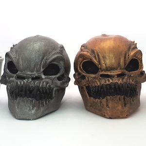 May include: Two decorative skull sculptures with menacing expressions. One is metallic gray, the other is bronze with gold highlights. Small potted cacti in white pots are arranged around the skulls, creating a unique and edgy home decor theme.
