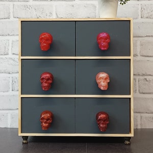 May include: A wooden chest of drawers with six drawers, each featuring a different coloured skull-shaped drawer pull. The drawers are dark grey and the chest has a light wood trim. The chest is standing on a black floor.