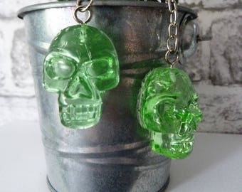 May include: Two green glass skull keychains hanging from silver chains. The keychains are hanging from a metal bucket with a green succulent plant inside.