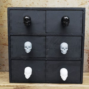 May include: A black wooden dresser with six drawers. Each drawer has a skull-shaped knob. The top two drawers have black skull knobs, the middle two drawers have gray skull knobs, and the bottom two drawers have white skull knobs.