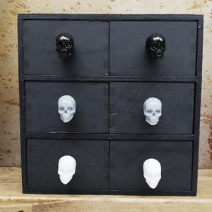 May include: Black wooden drawer unit with six drawers. Each drawer has a skull-shaped knob. The top two drawers have black skull knobs, the middle two drawers have gray skull knobs, and the bottom two drawers have white skull knobs.