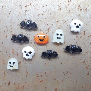 Puede incluir: Una colección de adornos de resina con temática de Halloween, que incluyen murciélagos, fantasmas, una calabaza y una calavera. Los adornos son blancos, negros y naranjas con detalles de purpurina.