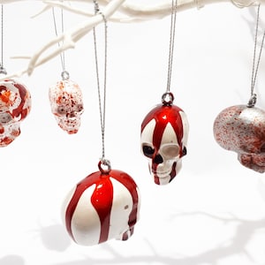 May include: Five white skull ornaments with red paint splatter and drips hanging from a white branch. The ornaments are a Halloween decoration.