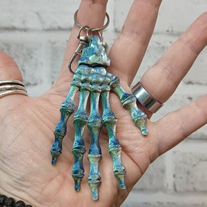 May include: A blue and green skeleton hand keyring with a silver ring. The keyring is held in a hand.