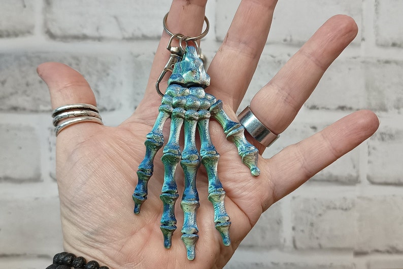 May include: A blue and green skeleton hand keyring with a silver ring. The keyring is held in a hand.