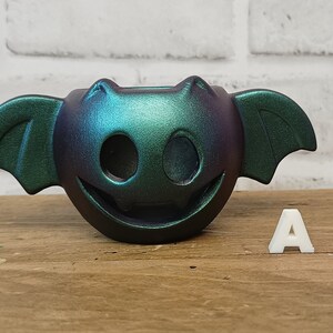 May include: A teal and purple iridescent ceramic bat-shaped planter with a smiling face and two holes for eyes. The planter is sitting on a wooden surface with a white letter A next to it.
