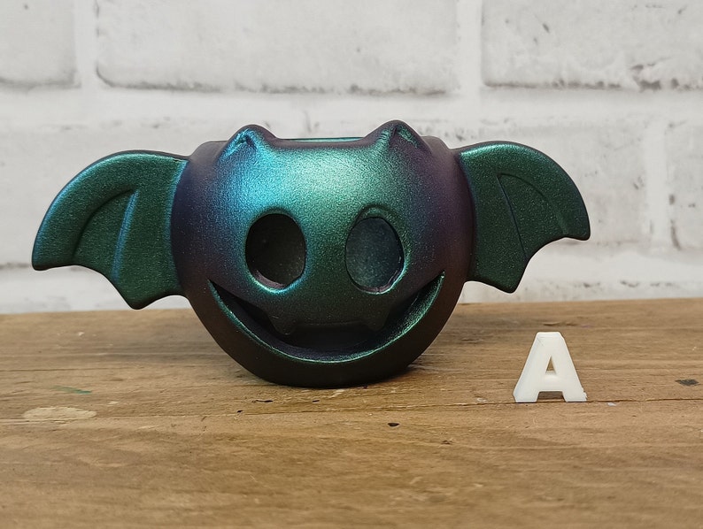 May include: A teal and purple iridescent ceramic bat-shaped planter with a smiling face and two holes for eyes. The planter is sitting on a wooden surface with a white letter A next to it.