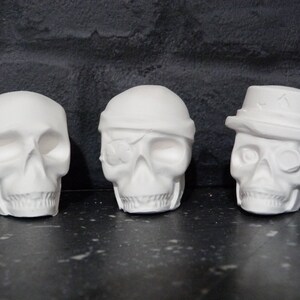May include: Three white plaster skull figurines. One skull is wearing a hat, another has an eye patch, and the third is a simple skull.