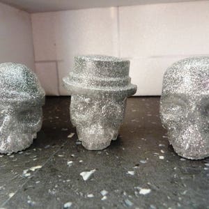 May include: Three silver glitter skull-shaped ornaments. The skull on the left has a hat. The skull in the middle has a hat with a brim. The skull on the right has no hat.