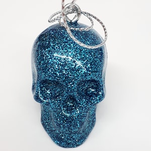 Puede incluir: Un adorno de calavera azul con purpurina y una cuerda plateada para colgar.